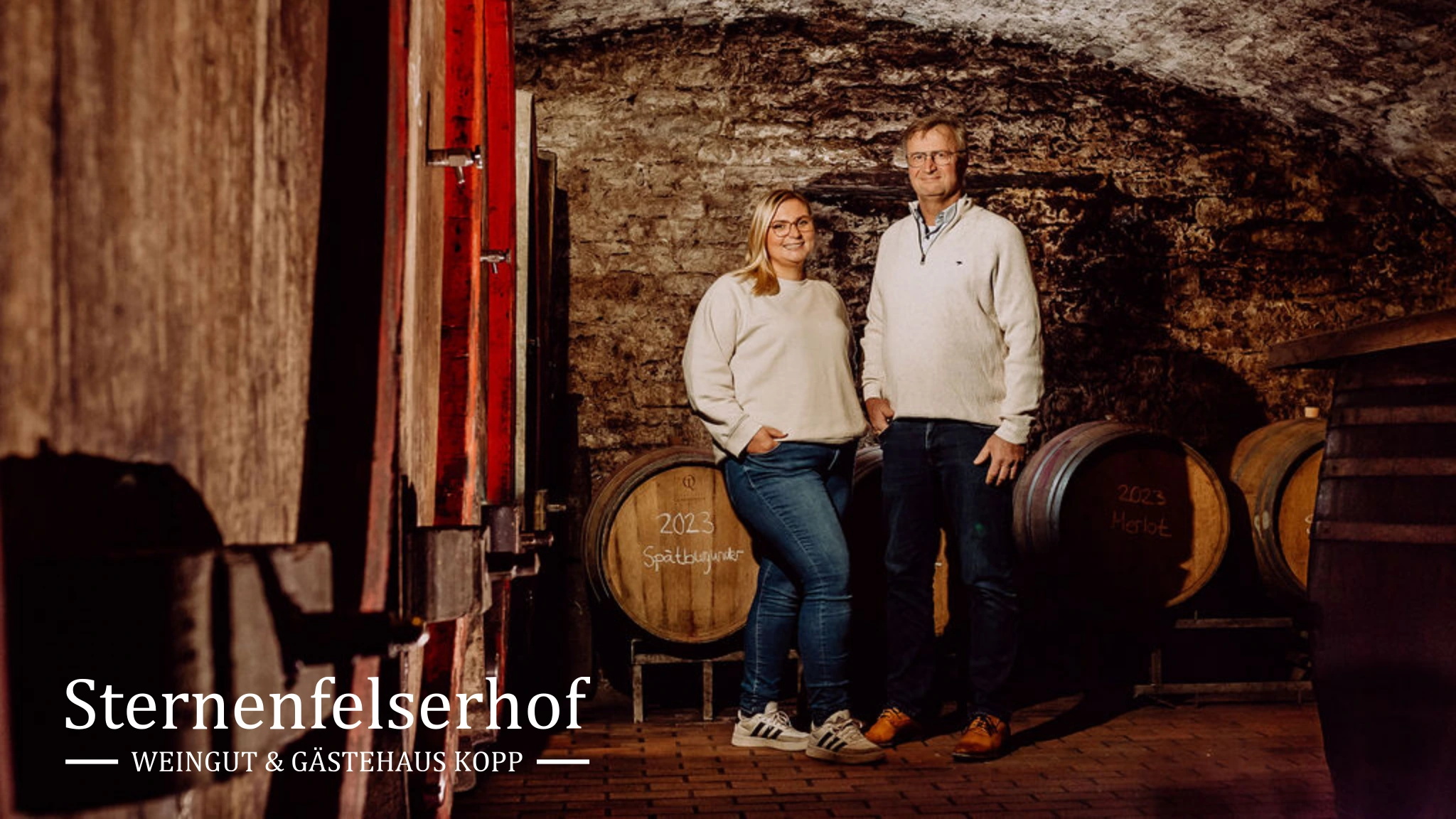 Jana und Rudolf Kopp im Holzfasskeller ihres Weinguts in Nierstein. Unten links steht das weiße Sternenfelserhof-Logo.